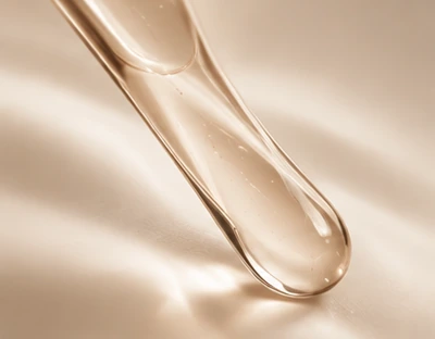 A clear serum drop at the end of a glass pipette, on a soft champagne background.